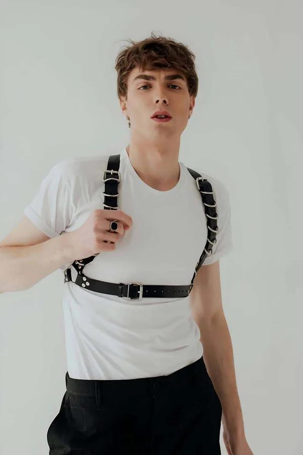 Black faux leather chest harness with silver O-rings on shoulder straps and rectangular buckle waist band worn over white t-s