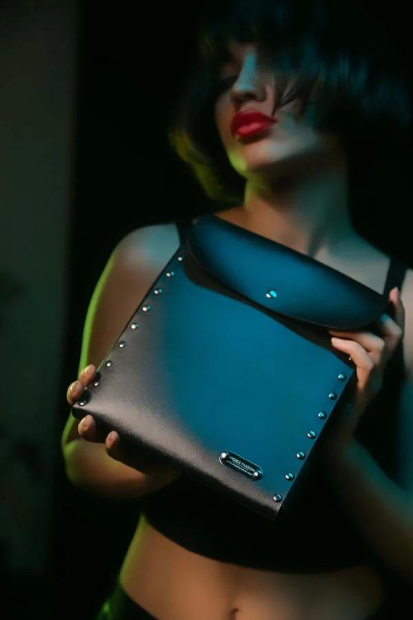Black faux leather shoulder bag with silver edge studs and metal branding plate, structured rectangular shape